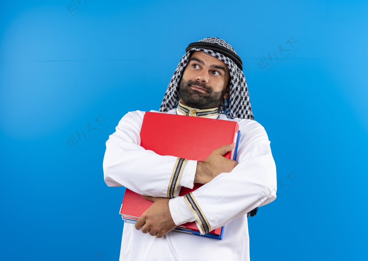 困惑身着传统服装的阿拉伯商人拿着两个文件夹 站在蓝色的墙上困惑地看着一边站在一边传统持有