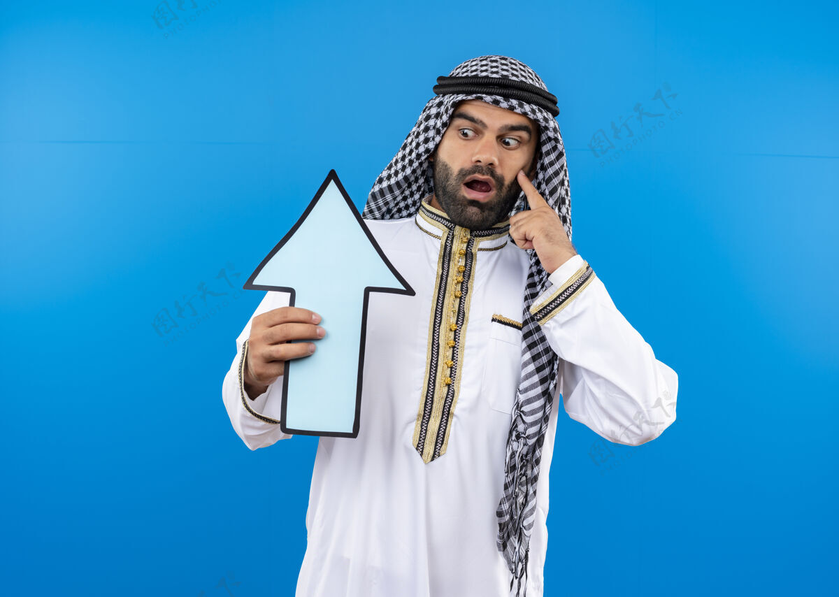 惊讶身着传统服饰的阿拉伯男子手持蓝色的大箭头 站在蓝色的墙壁上惊奇地看着它传统拿着穿