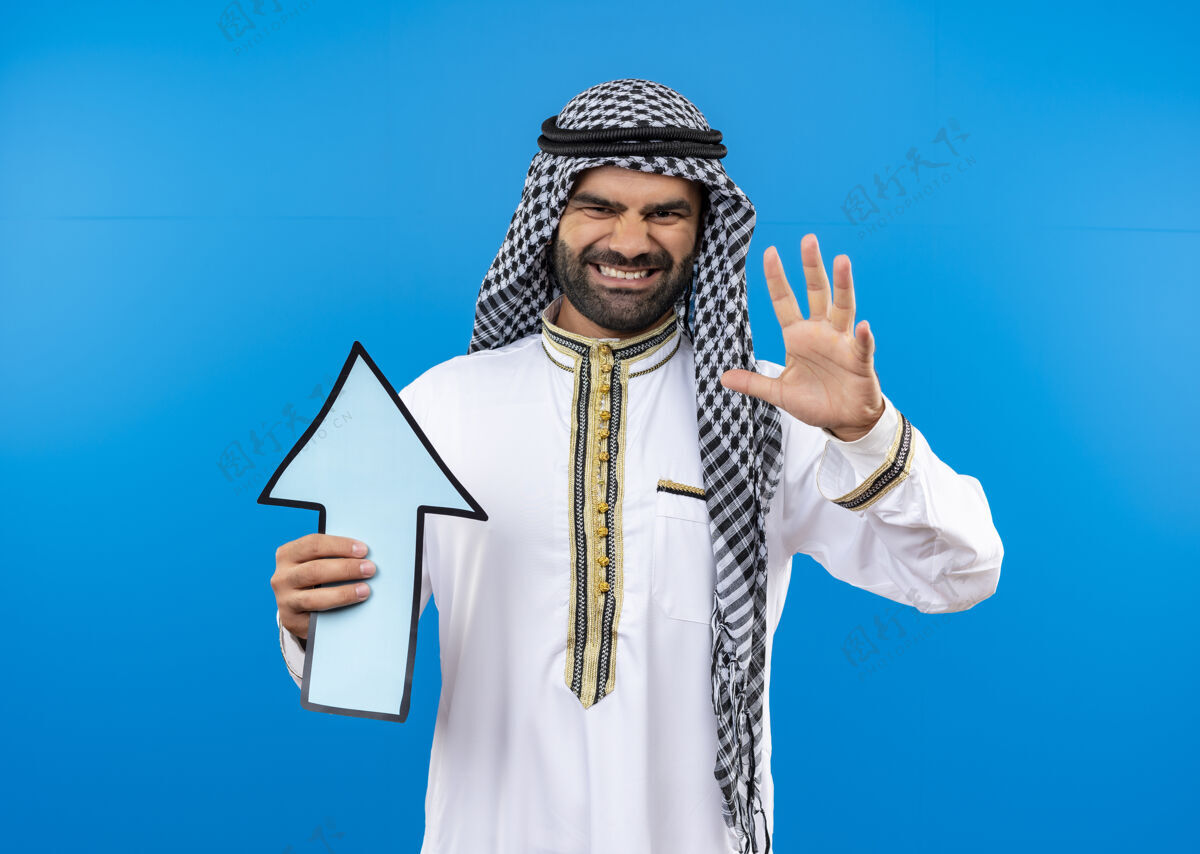 箭身着传统服装的阿拉伯男子手持蓝色大箭头 脸上带着恼怒的表情站在蓝色的墙上脸拿着人