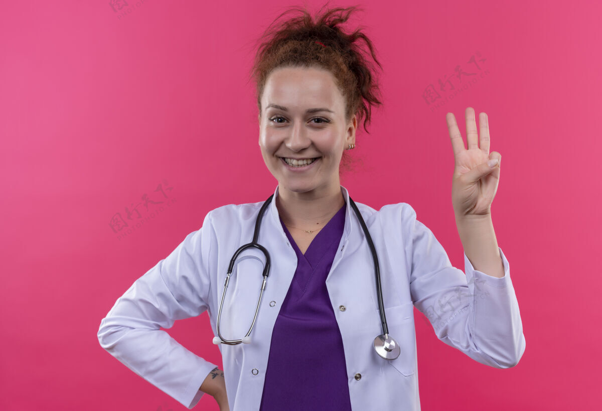 外套年轻的女医生 穿着白色外套 听诊器露出来 手指朝上 三号手指微笑着站在粉色的墙上指指点点号码医生