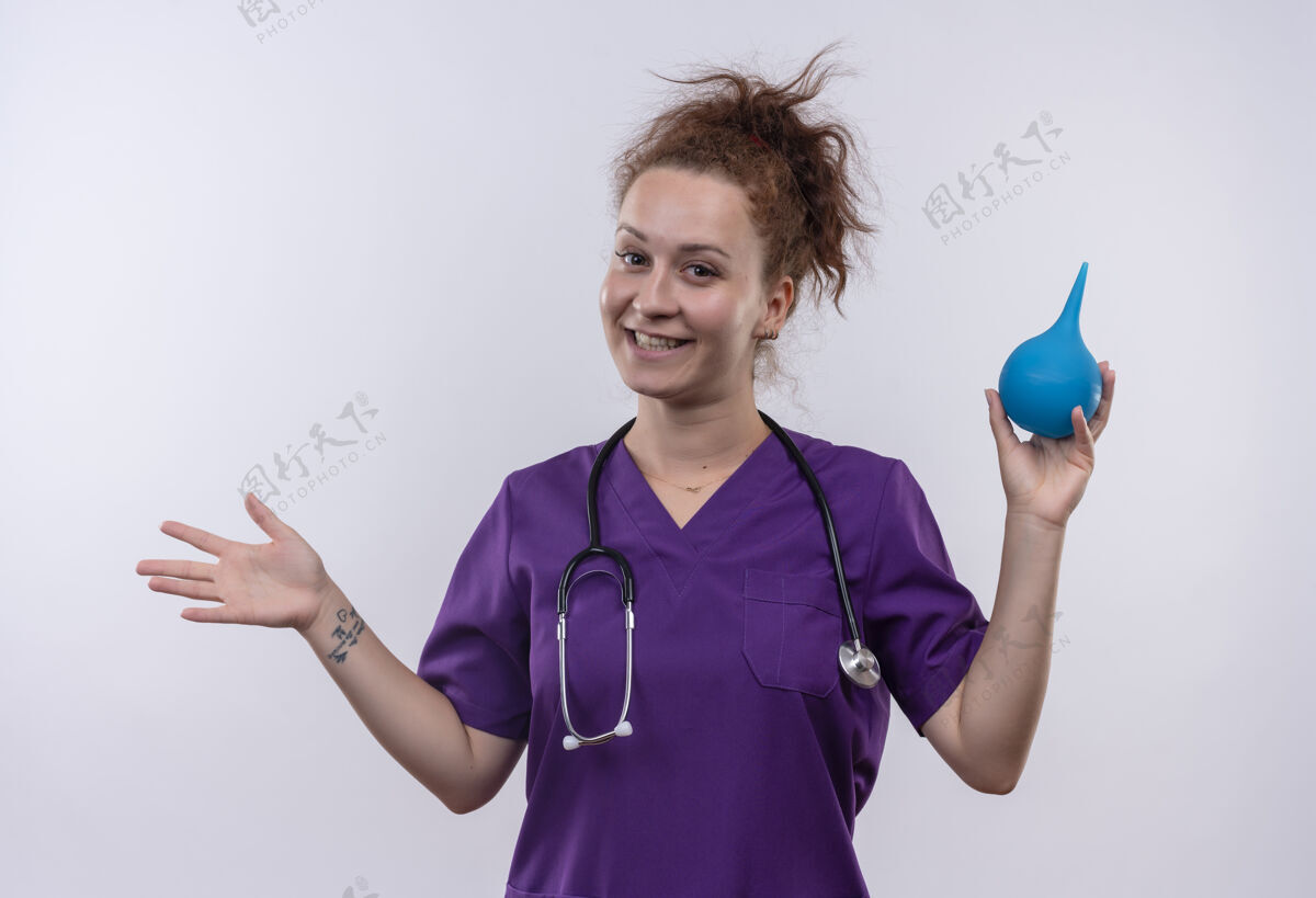 听诊器年轻女医生身穿医疗制服 手持听诊器 手持灌肠器 面带微笑 手臂站在白墙上女人微笑年轻人