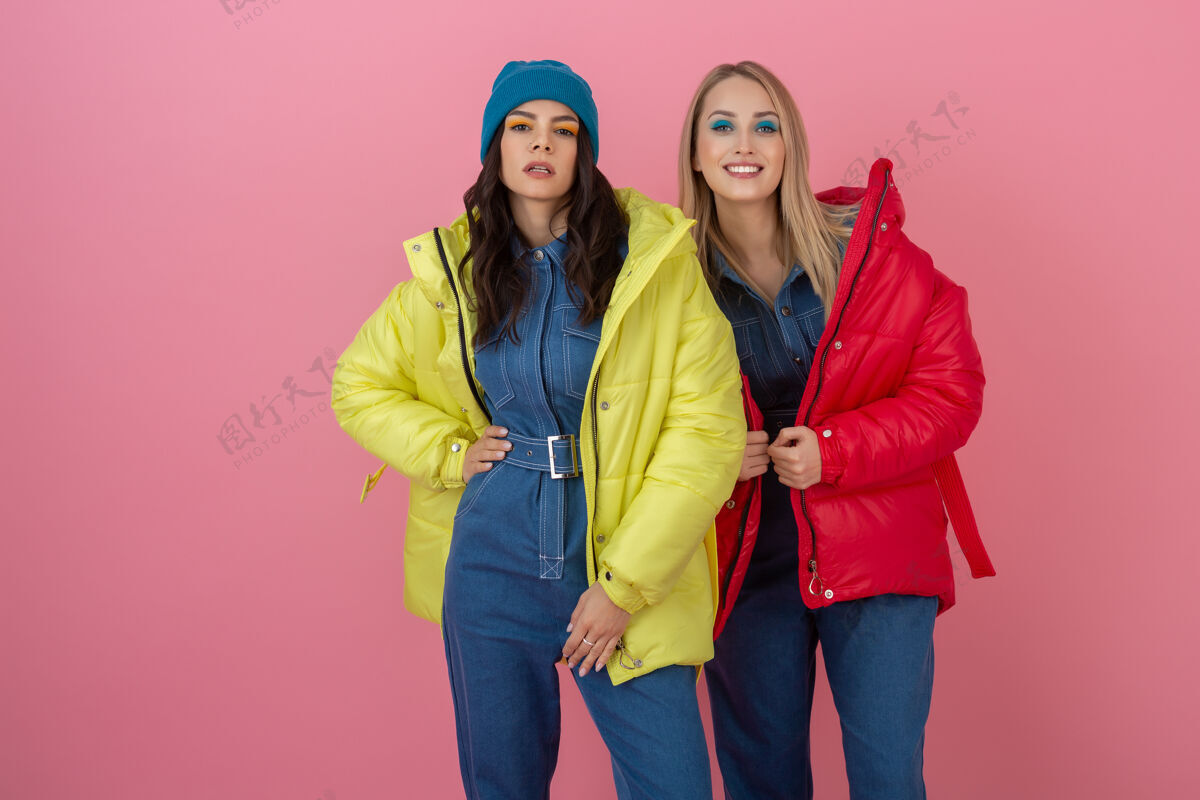 时尚两位兴奋迷人的女朋友活跃在粉色墙面上 身着鲜艳的红黄相间的冬季羽绒服 一起嬉戏 温暖外套时尚潮流模特人年轻