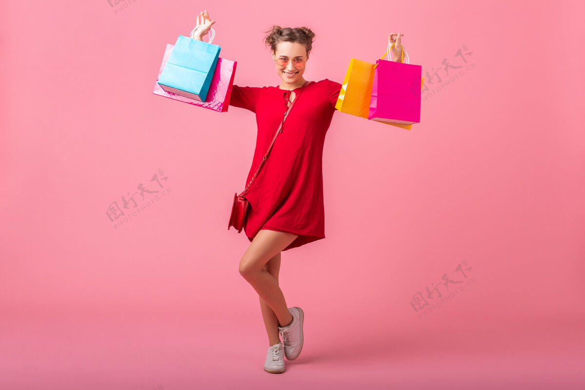 太阳镜迷人快乐微笑的时尚女性购物狂身着红色新潮连衣裙手持五颜六色的购物袋贴在粉色的墙上 销售兴奋 春夏时尚潮流顾客欢呼购买