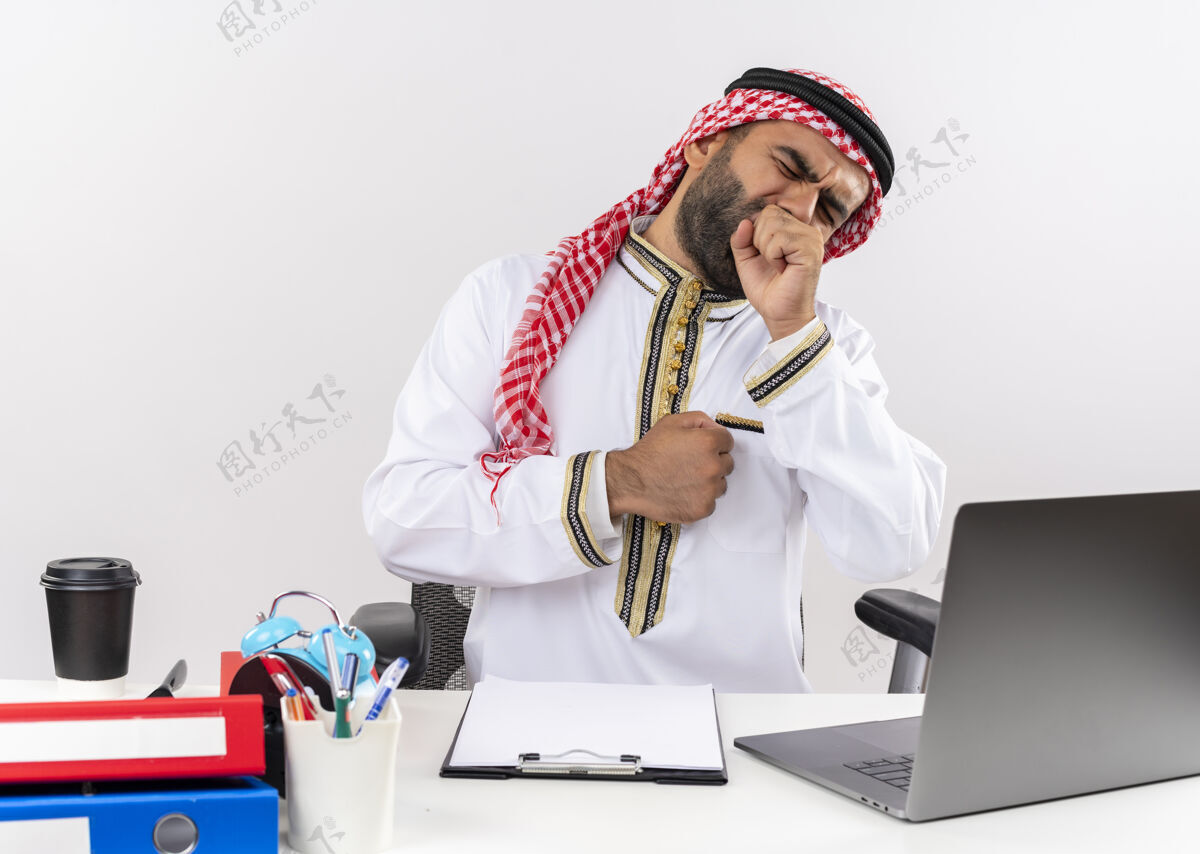 笔记本电脑身着传统服装的阿拉伯商人坐在桌旁 手提电脑看起来很疲惫 打着哈欠在办公室工作累穿阿拉伯语