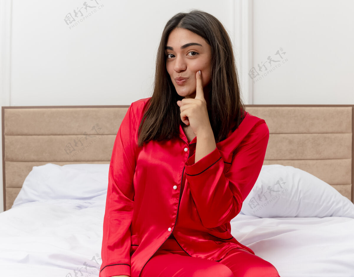 相机穿着红色睡衣在床上放松的年轻美女脸颊指着手指