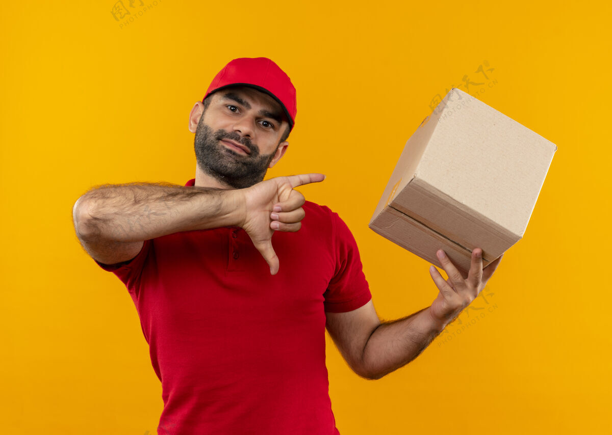 大拇指有胡子的送货员穿着红色制服 戴着帽子 拿着盒子 大拇指朝下 站在橙色的墙上展示放下站着