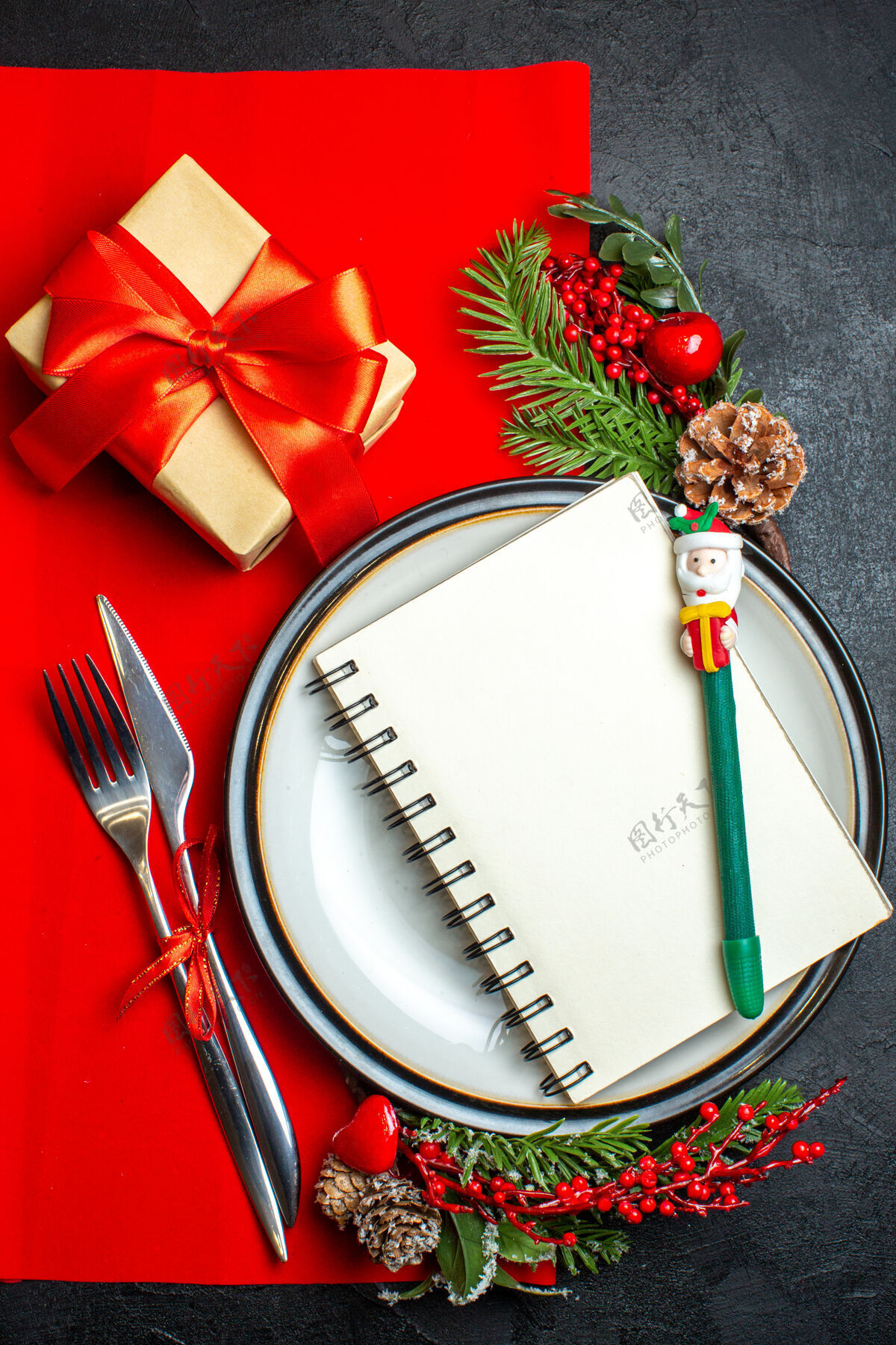 树枝新年背景的垂直视图用螺旋笔记本放在餐盘上餐具套装装饰配件杉木树枝旁边一张红色餐巾上的礼物餐盘圣诞笔记本