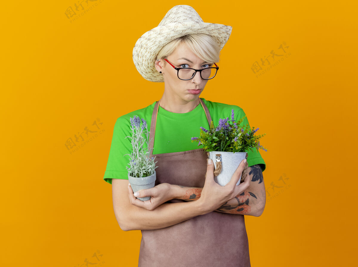 头发一个不满的年轻园丁 围裙上留着短发 戴着帽子 手里拿着盆栽植物 站在橙色背景下 皱着眉头看着摄像机植物围裙女人