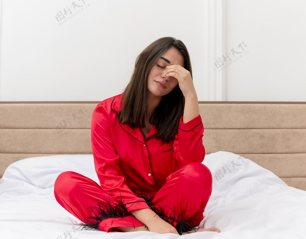 过度劳累穿着红色睡衣的年轻美女坐在床上 看起来很累 而且工作过度 她想在卧室内部的灯光背景下睡觉漂亮坐着睡眠