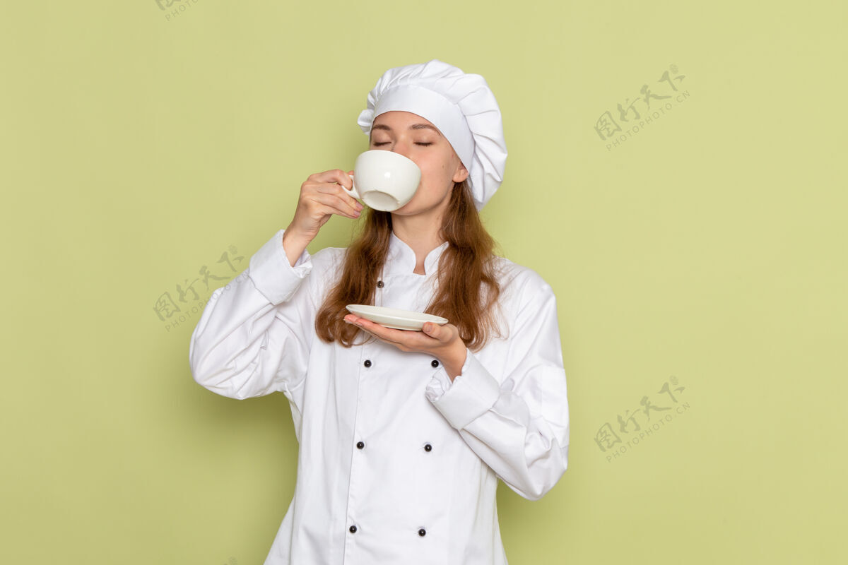 外套身着白色厨师服的女厨师在绿墙上喝咖啡的正面图饮用护士浴袍