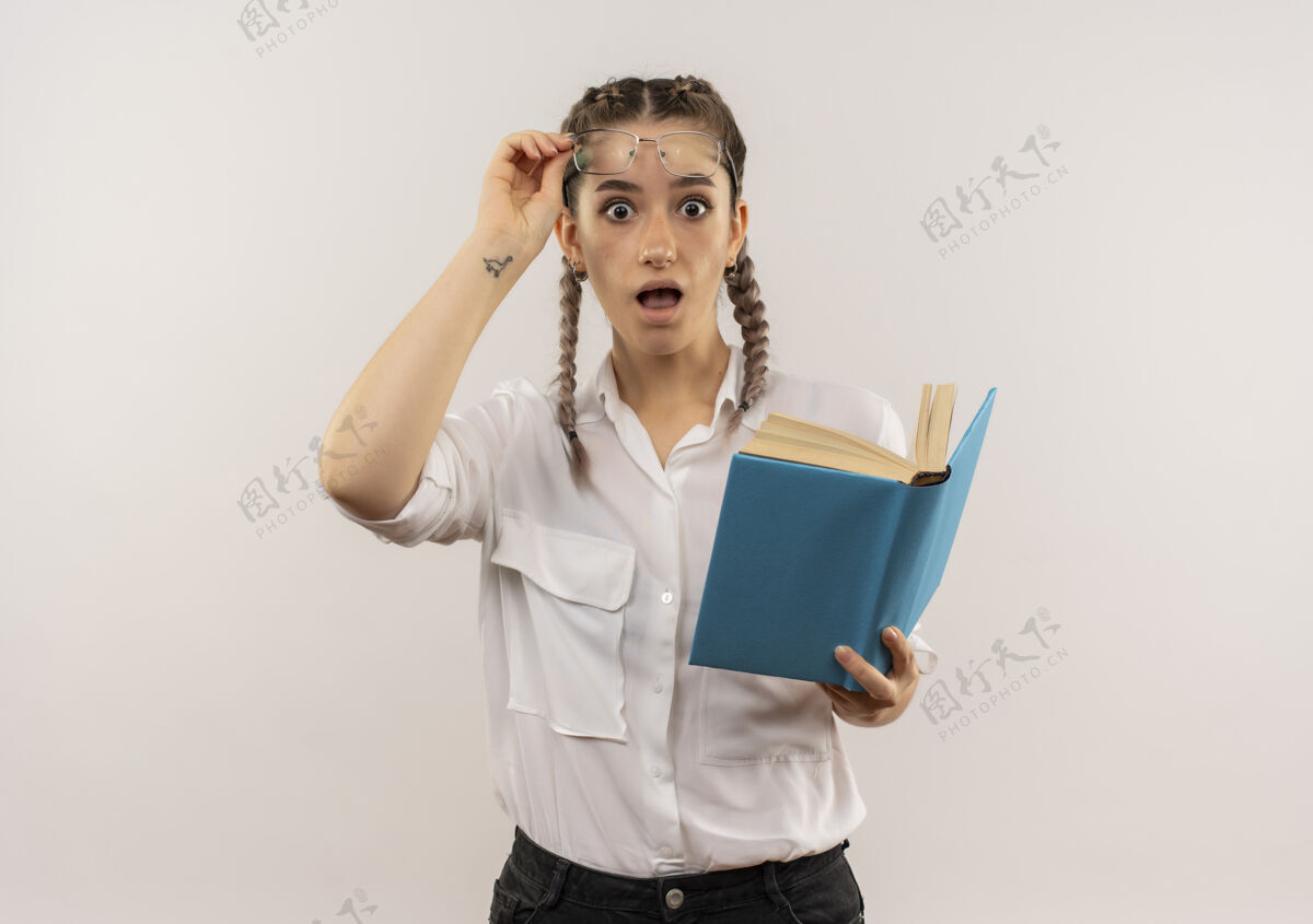 人戴着眼镜 梳着辫子 穿着白衬衫的年轻女学生手里拿着打开的书 望着前面摘下眼镜 站在白墙上显得很惊讶看书眼镜