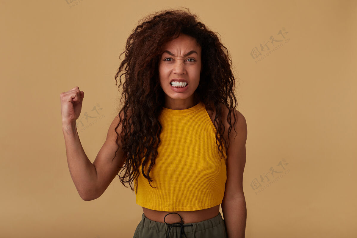愤怒摄影棚拍摄了一个恼怒的长发卷曲的黑皮肤女人在愤怒地看着镜头时做鬼脸 站在米色背景上不停地举起手女士长运动