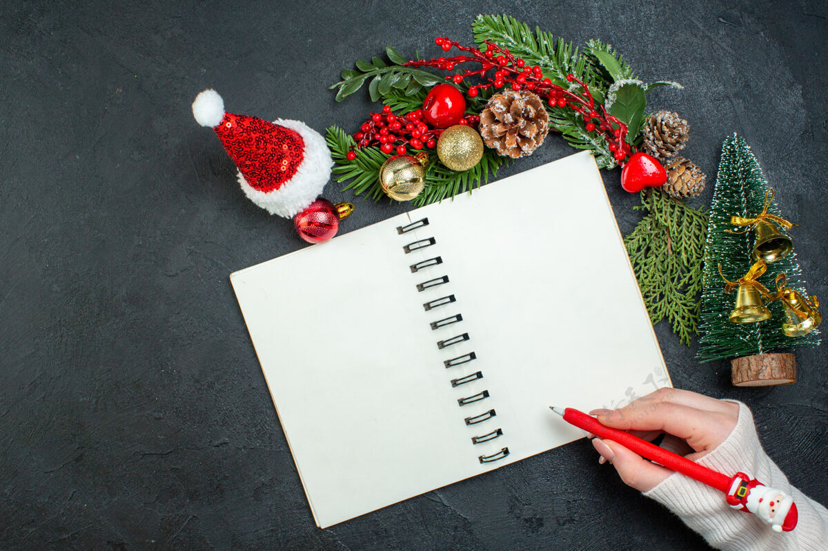 笔记本上图是圣诞心情与冷杉树枝圣诞树圣诞老人帽子手拿一支笔在黑色背景上的螺旋笔记本视图帽子圣诞老人