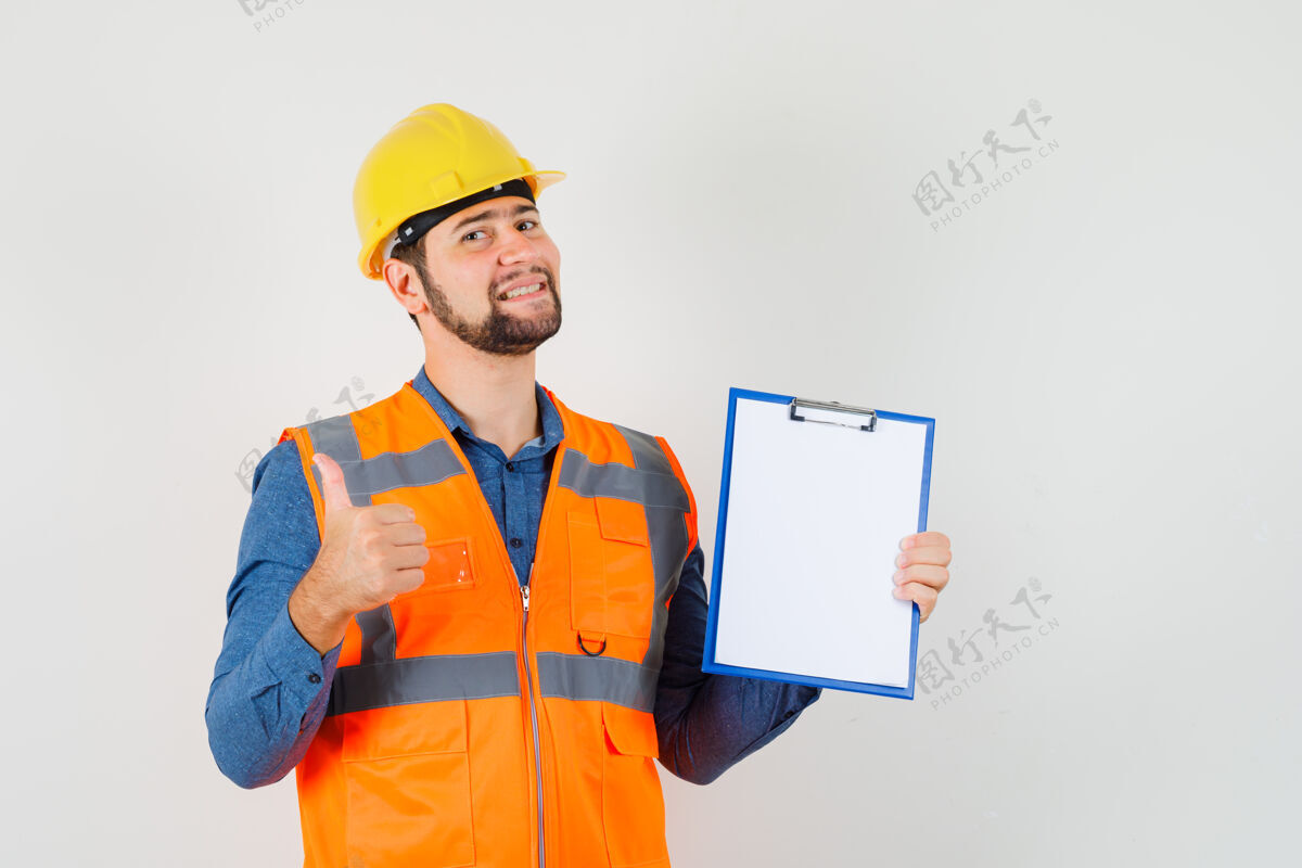 建筑商年轻的建筑工人穿着衬衫 背心 头盔 大拇指朝上 拿着剪贴板 看起来很高兴 正面视图建筑努力专业