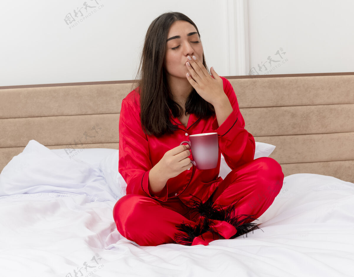 红色穿着红色睡衣的年轻美女坐在床上 一边喝着咖啡一边打呵欠 想在卧室内部的灯光背景下睡觉哈欠杯子女士
