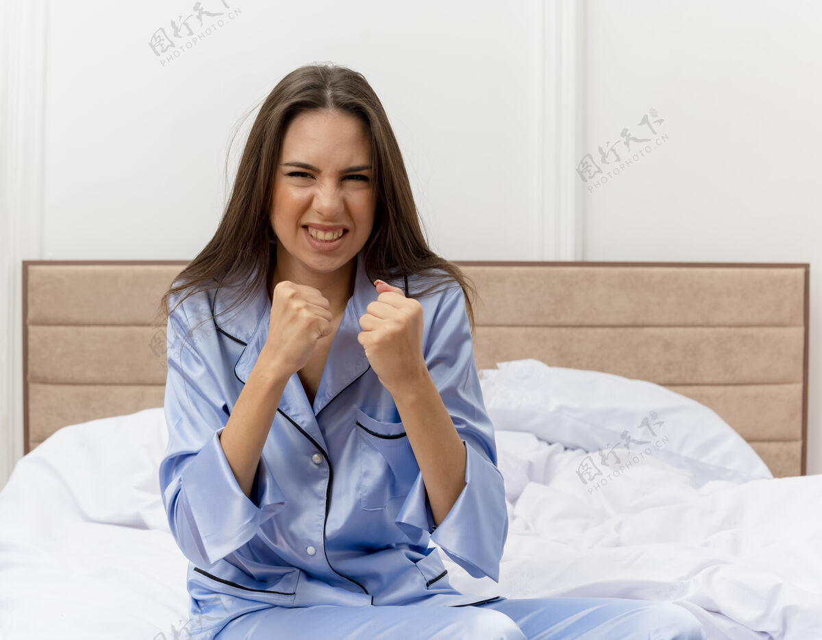 美丽穿着蓝色睡衣的年轻漂亮女人坐在床上看着相机握紧拳头愤怒的脸在卧室内部灯光背景下拳头床室内