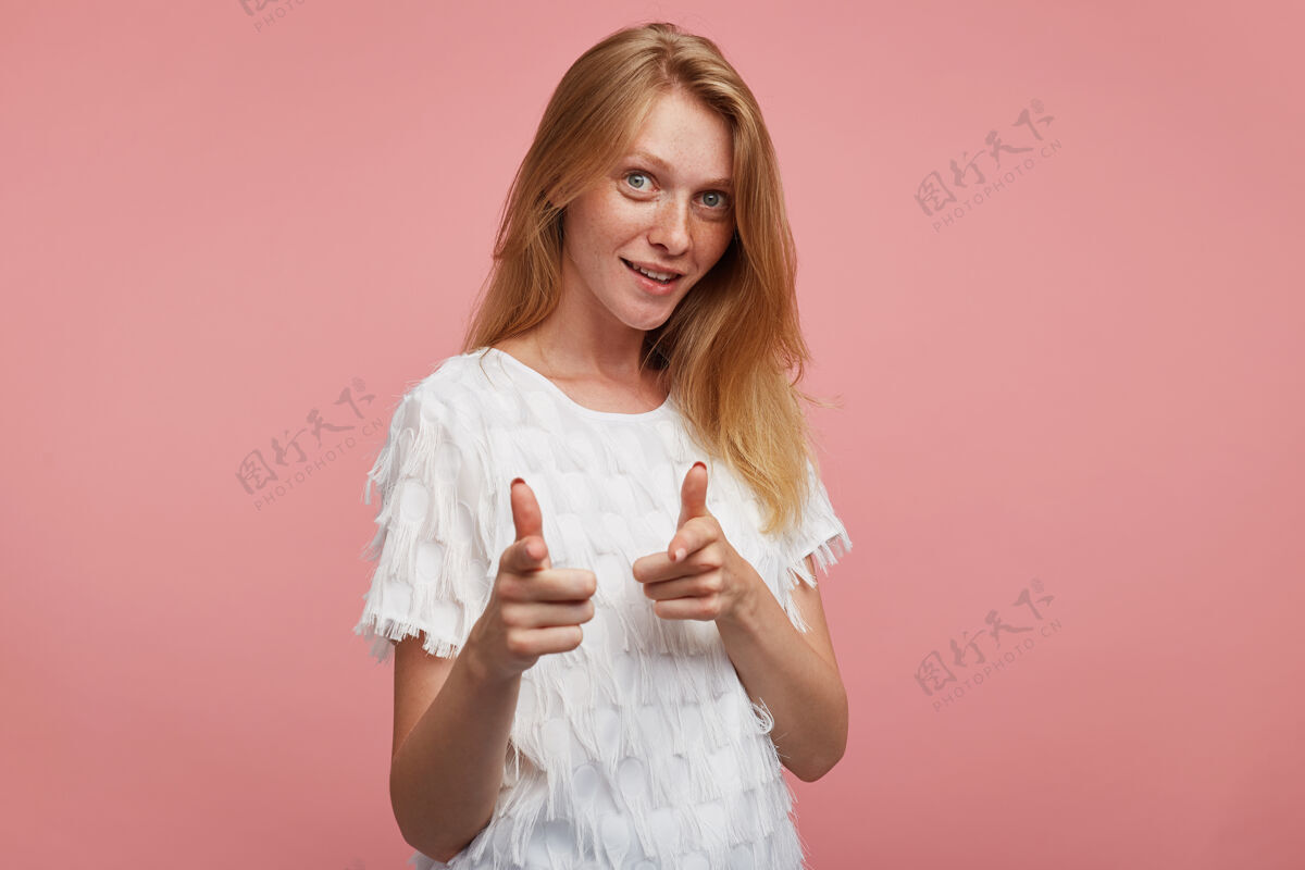 衬衫一幅正面的年轻漂亮红发女子的肖像 她有一双绿灰色的眼睛 站在粉色背景下 用食指对着镜头微微微笑红发抬高食指
