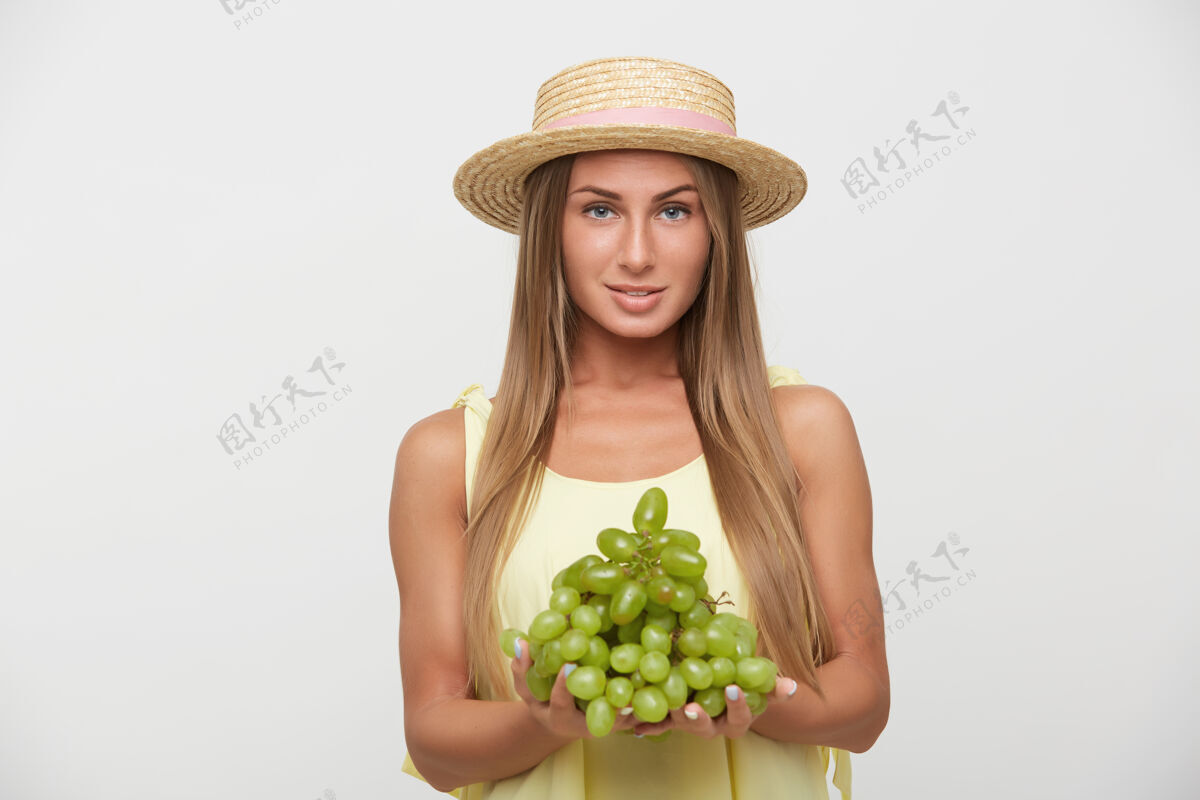 欢呼淡定的年轻迷人的金发女性 自然的妆容 带着淡淡的微笑积极地看着镜头 手持一大串绿色葡萄 在白色背景上摆姿势休闲女士年轻