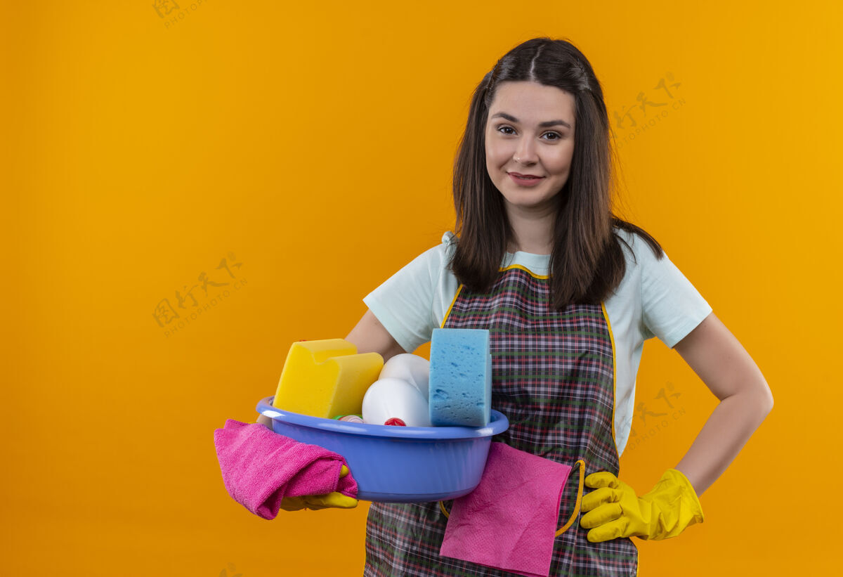 清洁年轻漂亮的女孩穿着围裙 戴着橡胶手套 拿着洗脸盆和清洁工具 友好地微笑着看着镜头橡皮女孩工具