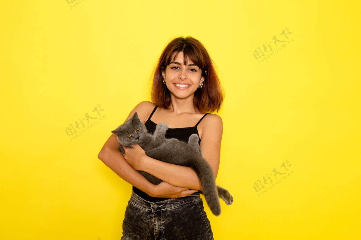 性感身穿黑色衬衫和灰色牛仔裤的年轻女性的正面视图 黄色墙上抱着可爱的灰色小猫女孩漂亮牛仔裤