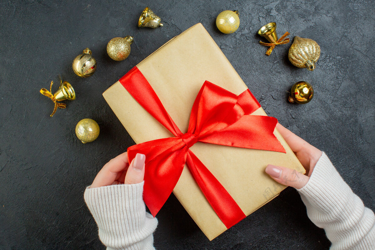 查看上图中 手拿着一个带有红色丝带和装饰配件的礼品盒 背景为深色圣诞节信封蝴蝶结