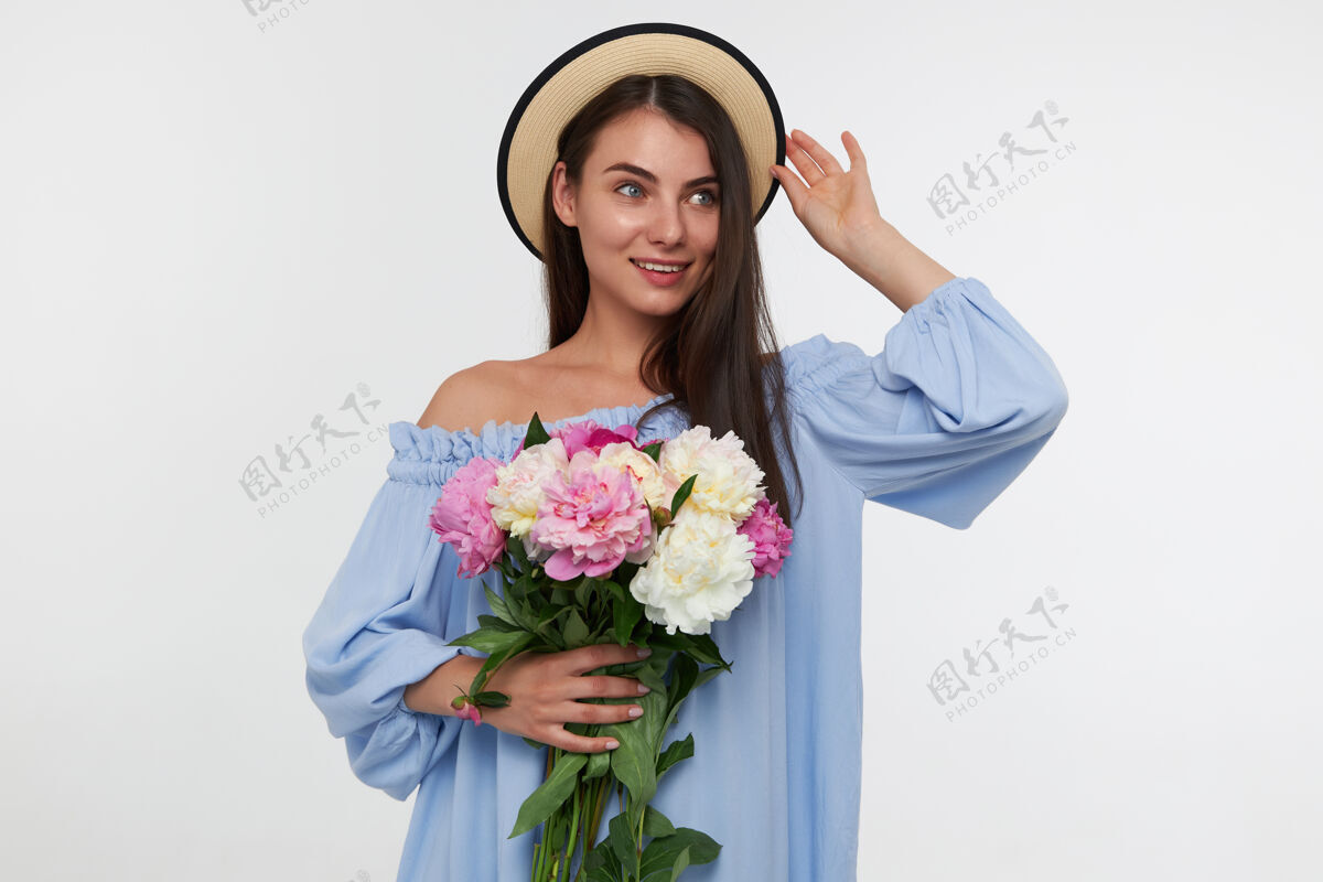 长年轻的女士 漂亮的女人 留着深褐色的长发 戴着一顶帽子和一件蓝色的裙子 手里拿着一束漂亮的花 摸着她的帽子 看着白色墙壁上的复制空间向右看调情服装微笑