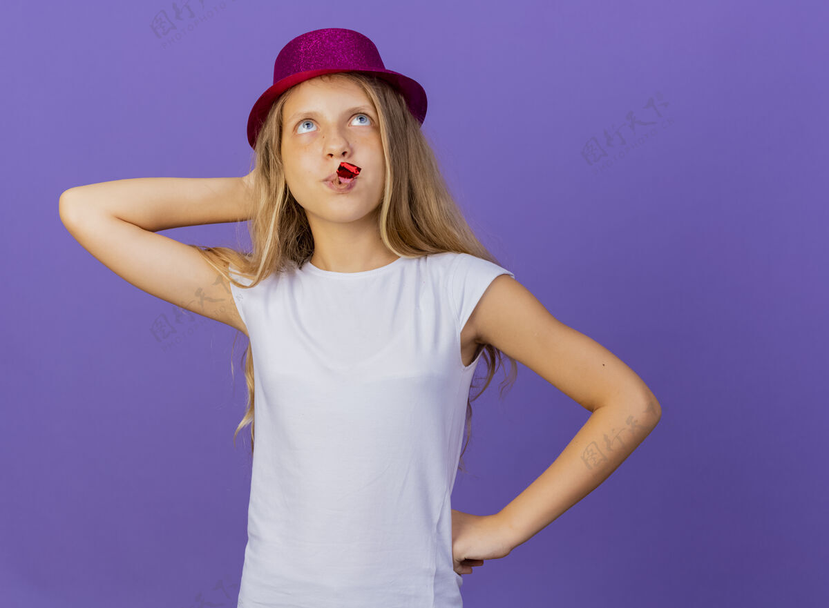 节日戴着节日帽子吹口哨的漂亮小女孩站在紫色的背景上 抬头看着困惑的生日派对概念小站哨子