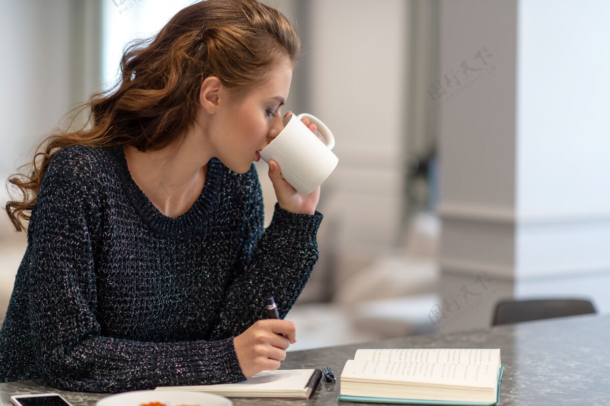女孩在家工作的年轻女子在厨房里用记事本她正在喝咖啡在家学习和工作的想法吸引房子家庭作业