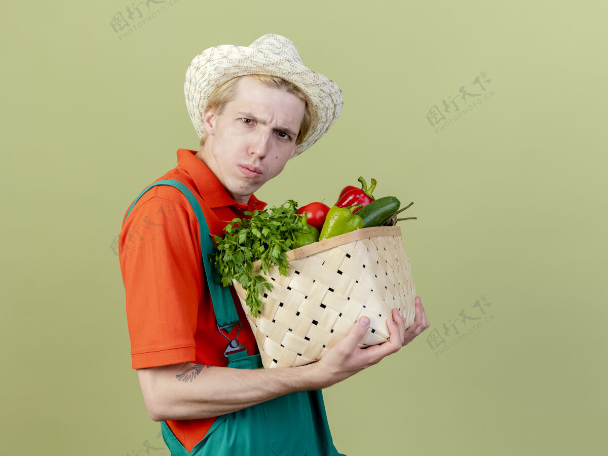 站着年轻的园丁穿着连体衣 戴着帽子 手里拿着装满蔬菜的箱子 站在灯光下严肃地皱着眉头看着摄像机皱眉板条箱男人