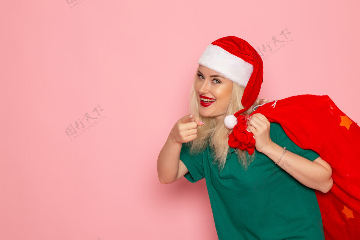 美丽正面图年轻女性提着红包带着礼物在粉色墙上度假模特圣诞新年彩照圣诞老人帽子风景圣诞老人