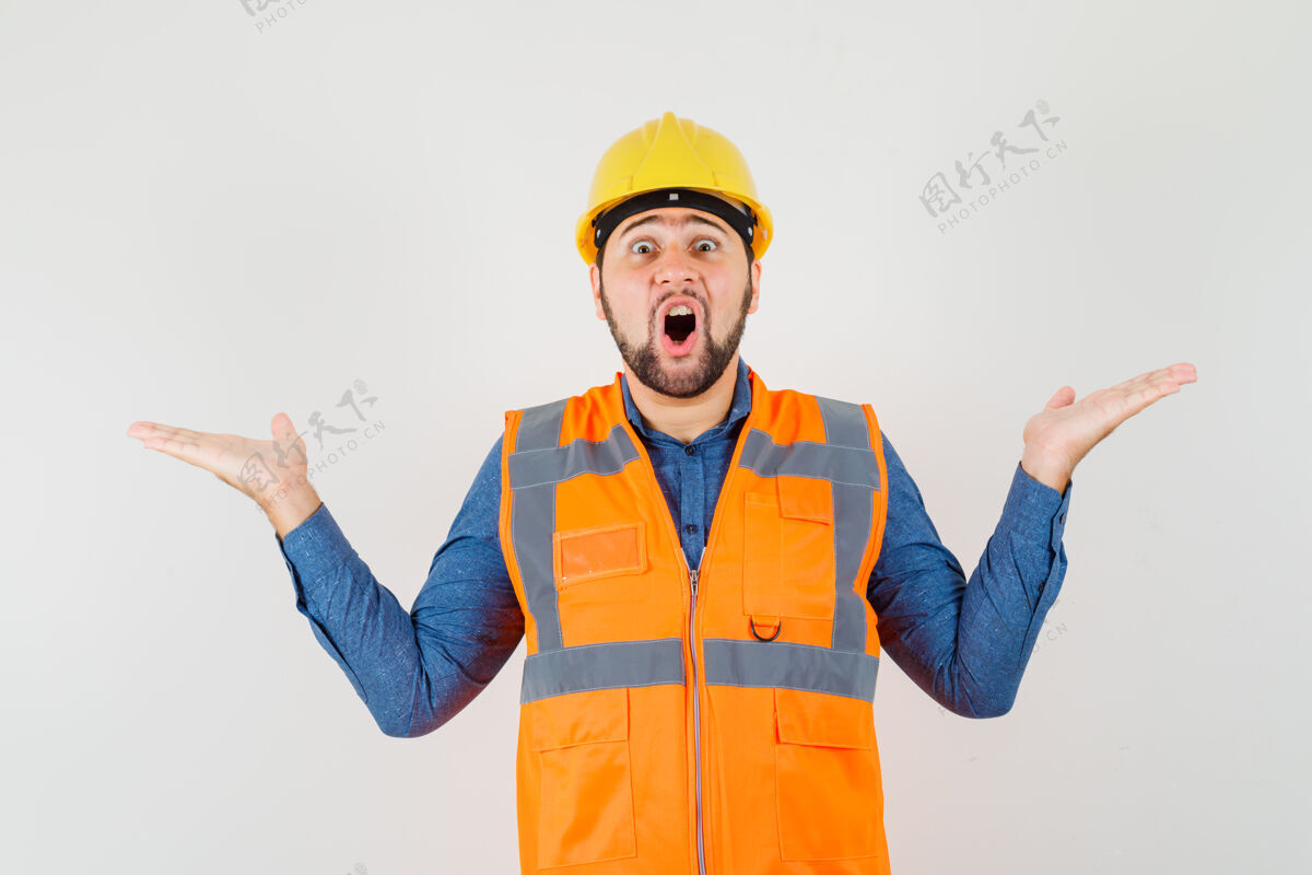 前面年轻的建设者在衬衫 背心 头盔上做着秤的手势 看上去很惊讶 正面视图工具建筑物勤杂工