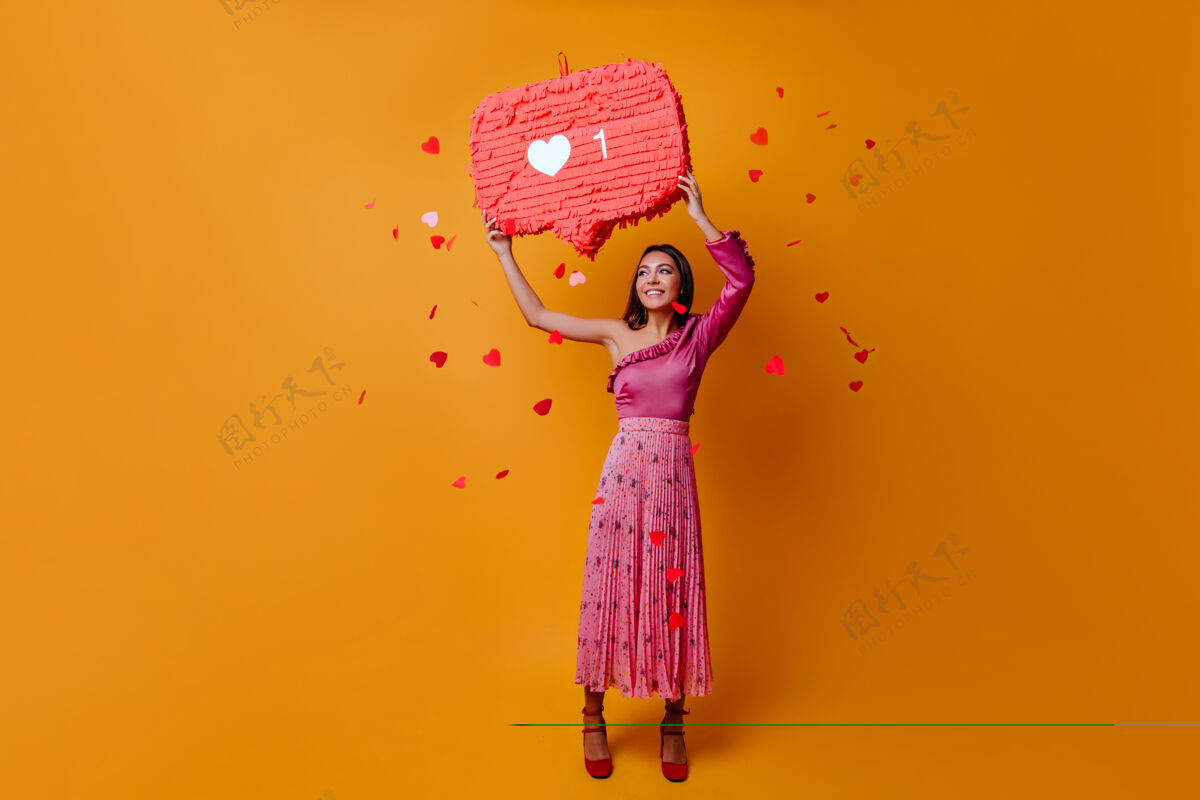 连接23岁的幸运迷人的女人拿着instagram的like标志 在橙色的墙上摆满了五彩纸屑喜欢信息舞蹈