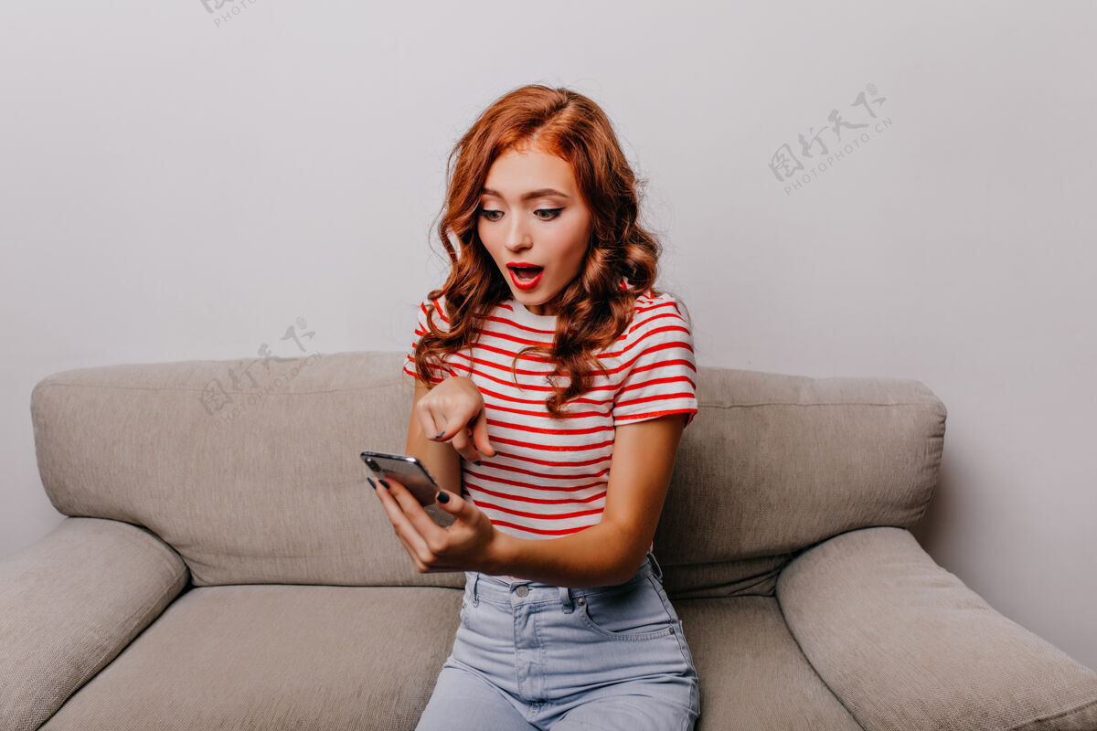 室内坐在沙发上惊讶地看着手机屏幕的女士穿着条纹t恤的姜汁女孩和智能手机合影红发漂亮情感
