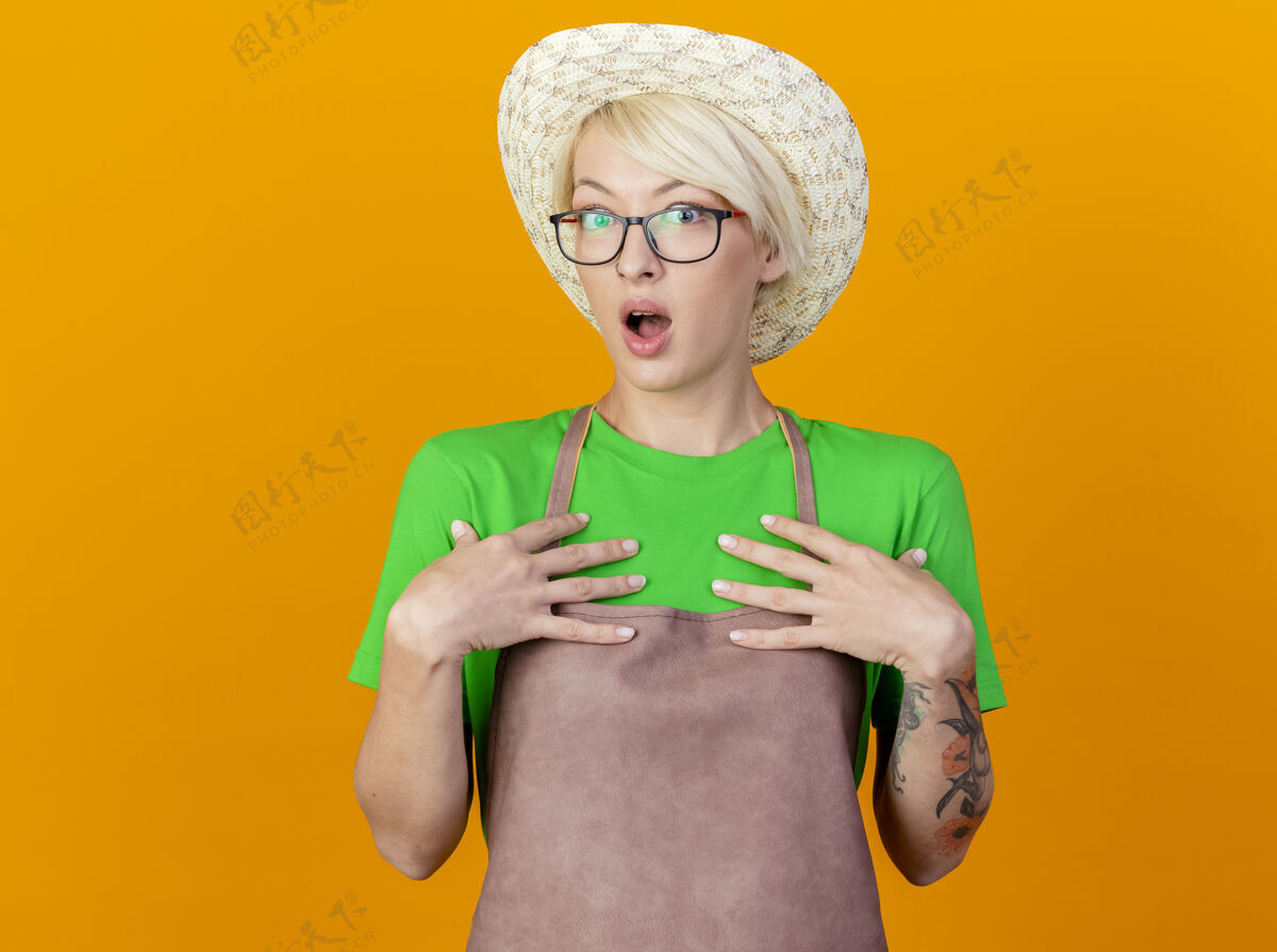 她自己穿着围裙 戴着帽子 留着短发的年轻园丁女人看着自己站在橙色背景下惊讶地看着摄像机围裙指向短裤