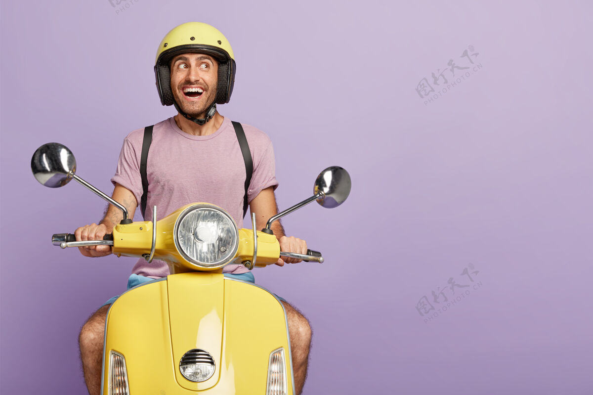 满意快乐的男性车手或信使开着黄色的踏板车 戴着防护头盔 穿着休闲t恤 在自己的交通工具上摆姿势 高兴地看着旁边 交通工具 隔离在紫色的墙上 空白处积极运输休闲