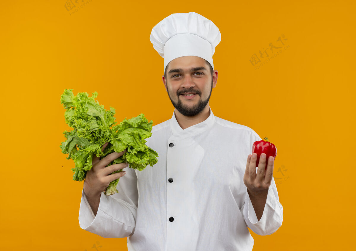 年轻年轻的男厨师身着厨师制服 手里拿着辣椒和莴苣隔离在橙色的空间里制服拿着生菜