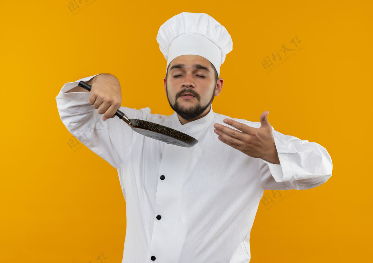 年轻身着厨师制服的年轻男厨师手拿煎锅 闭着眼睛闻着 手放在橙色空间的空气中制服嗅眼睛