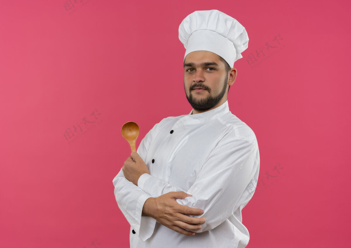 制服高兴的年轻男厨师穿着厨师制服拿着勺子 把手放在手肘上孤立的粉红色空间手空间粉红色