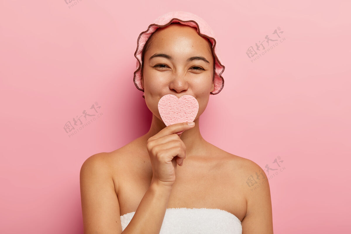 光滑美丽满足的韩国女性抱着心形海绵 洗脸 清洁皮肤 呵护肤色 做美容spa 裹着毛巾站在浴室里 戴着浴帽包装洗漱面部