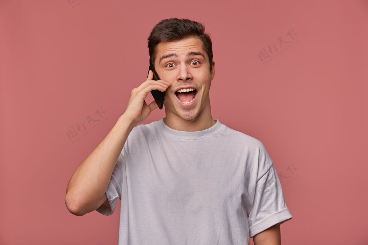 惊喜照片中的年轻人穿着一件白t恤 打电话 听到一个难以置信的消息 站在粉红色的地板上 张大嘴巴 表情震惊宽难以置信帅哥
