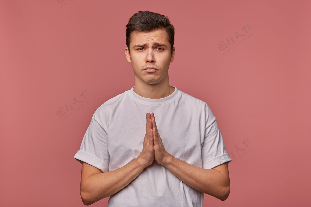 悲伤一幅年轻悲伤的人的肖像 穿着一件空白的t恤 希望好运 做着祈祷的手势 站在粉色的地面上 带着不开心的表情不快男孩年轻