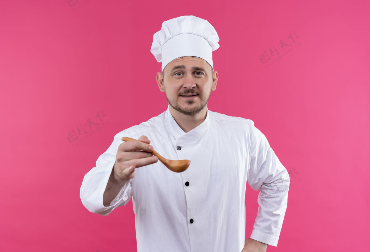 年轻高兴的年轻帅哥厨师穿着厨师制服伸出汤匙孤立在粉红色的空间请粉红色烹饪