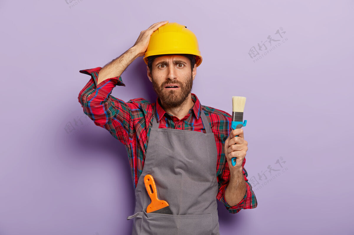 头盔失望的工业工人戴着安全帽 穿着休闲制服 拿着画笔作画 是专业画家 有着不悦的表情 隔着紫色的墙油漆不高兴重建