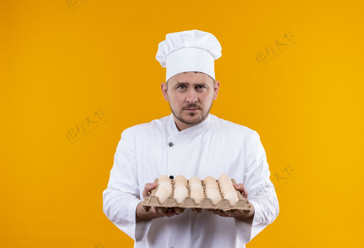 英俊年轻帅气的厨师穿着厨师制服 手里拿着一盒鸡蛋 看起来孤立在橙色的空间里橙色持有鸡蛋