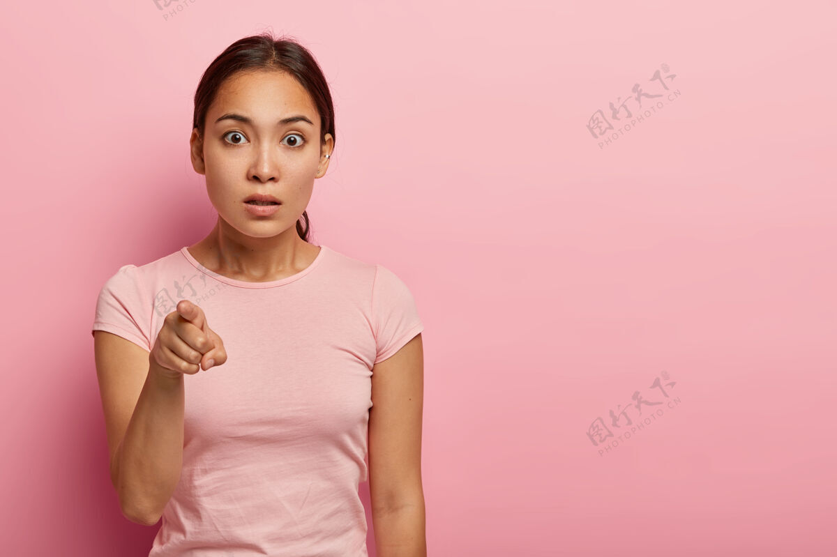 人惊艳的亚洲女人横拍指着食指 不好意思注意到什么奇怪的东西 有一头乌黑的头发梳在马尾上 穿着玫瑰色的t恤 孤立在粉色的墙上恐惧震惊害怕