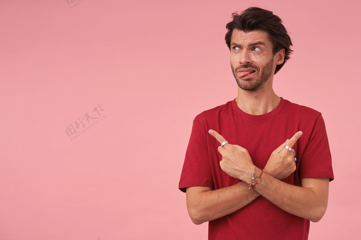 情绪一幅年轻人的画像 他留着时髦的发型 用食指指向不同的方向 站在粉色的红色t恤上 一边看一边露出舌头发型摆姿势指数