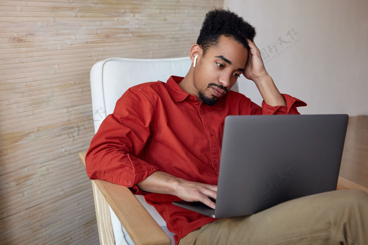 年轻疲惫的年轻卷曲的深色皮肤的男性 留着胡须 手举着头 专注地看着笔记本电脑的屏幕 坐在家里的椅子上室内非洲头