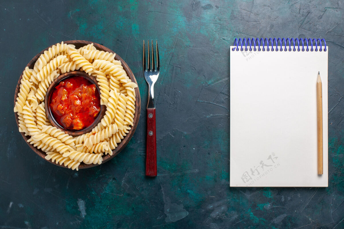 面团顶视图形状意大利面食与番茄酱和记事本上的深蓝色背景意大利酱菜西红柿