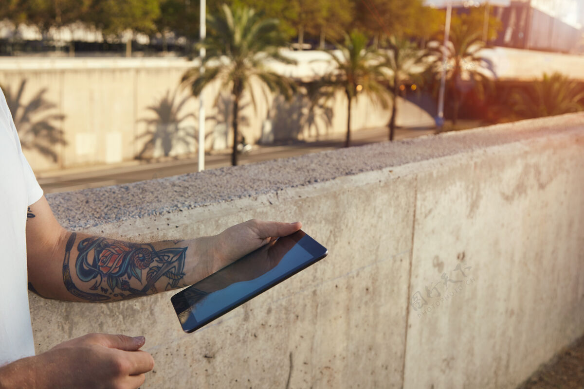 数字平板电脑一个纹身的男人拿着一块黑色的石碑站在一堵灰色的水泥墙旁边 城市里有棕榈树太阳外观企业