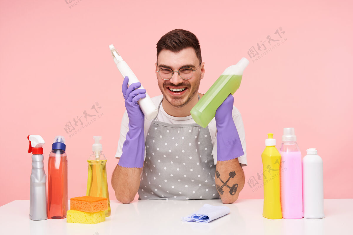 男人室内写真：一个快乐的年轻漂亮的短发黑发男人 手里拿着几瓶家用化学品 坐在粉红色的椅子上开心地笑着抬高清洁室内
